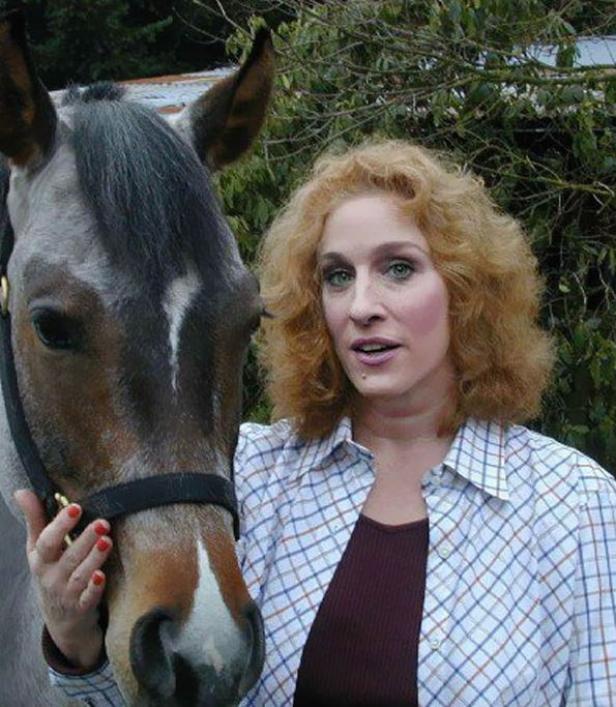 Eine Frau mit lockigem, rötlichem Haar steht neben einem grauen Pferd.