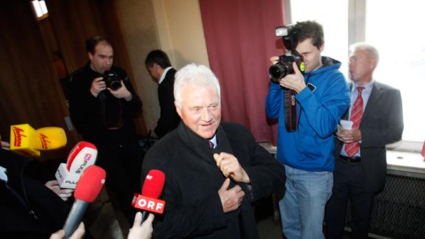 Ein Mann im schwarzen Mantel wird von Reportern und Fotografen umringt.