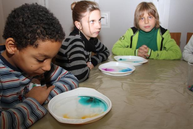 Drei Kinder sitzen an einem Tisch und experimentieren mit Farben in Wasserschalen.