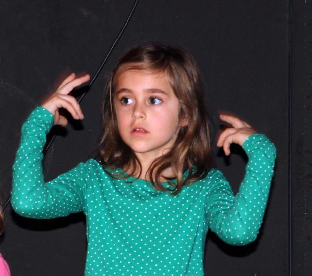 Ein junges Mädchen mit einem grün-weiß gepunkteten Oberteil gestikuliert vor einem dunklen Hintergrund.