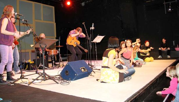 Eine Band spielt auf einer Bühne vor einem sitzenden Publikum aus Kindern.
