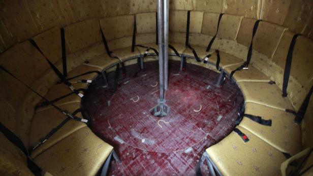 Ein runder Raum mit gepolsterten Sitzen, Sicherheitsgurten und einer Stange in der Mitte.