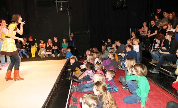 Eine Frau in einem gelben Kleid steht vor einer Gruppe von Kindern auf einer Bühne.