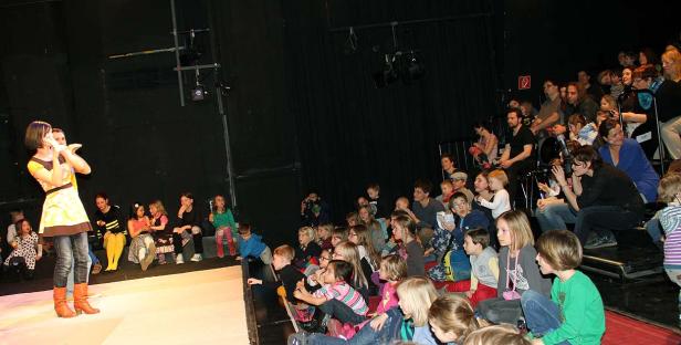 Eine Frau steht auf einer Bühne vor einem Publikum aus Kindern und Erwachsenen.