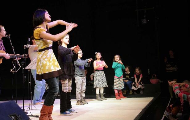 Eine Frau leitet eine Gruppe Kinder auf einer Bühne an, während ein Mann Gitarre spielt.
