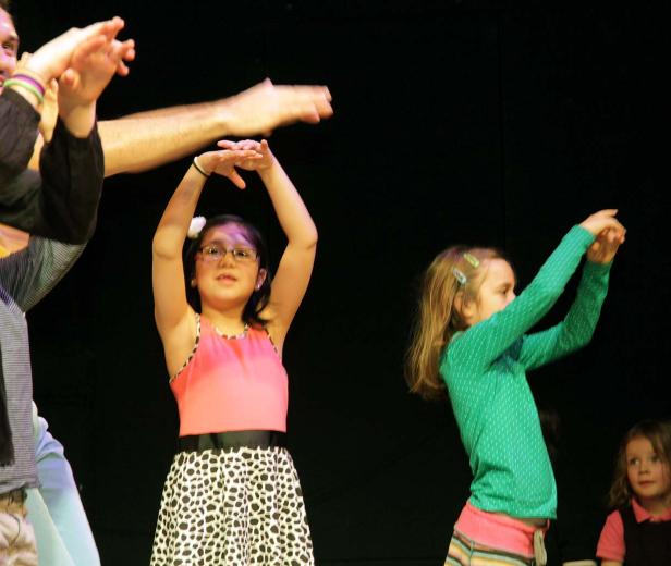 Mehrere Kinder und ein Erwachsener tanzen auf einer Bühne vor einem schwarzen Hintergrund.