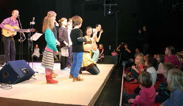 Eine Gruppe Kinder steht mit ihren Betreuern auf einer Bühne, während ein Mann Gitarre spielt und singt. Eine Zuschauergruppe sitzt davor.
