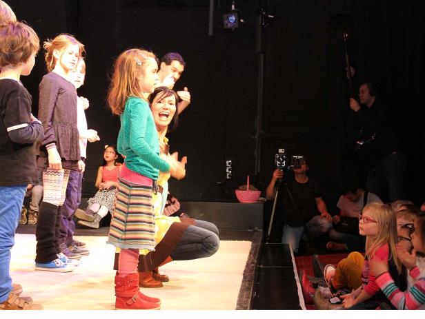 Eine Gruppe Kinder steht auf einer Bühne, während eine Frau sie anfeuert.