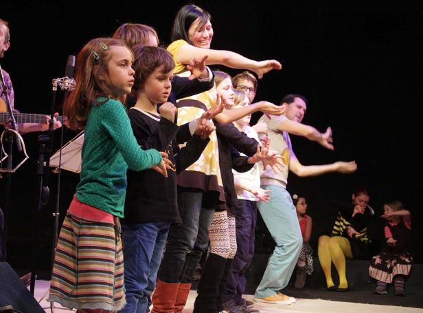 Eine Gruppe von Kindern und Erwachsenen steht auf einer Bühne und führt eine synchrone Bewegung auf.