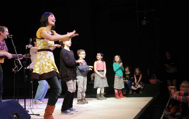 Eine Frau singt mit einer Gruppe von Kindern auf einer Bühne.