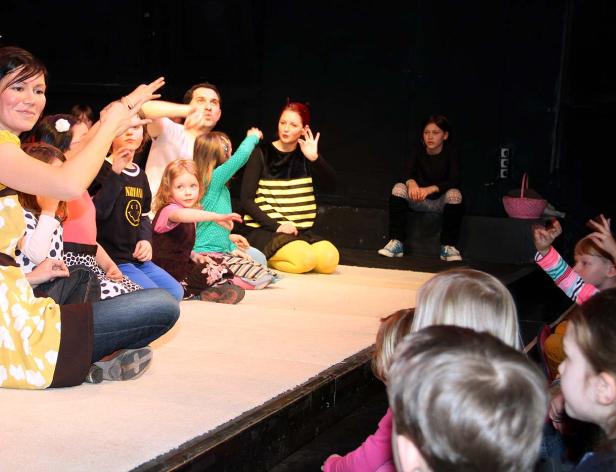 Eine Gruppe von Kindern sitzt auf einer Bühne, einige mit erhobenen Händen.