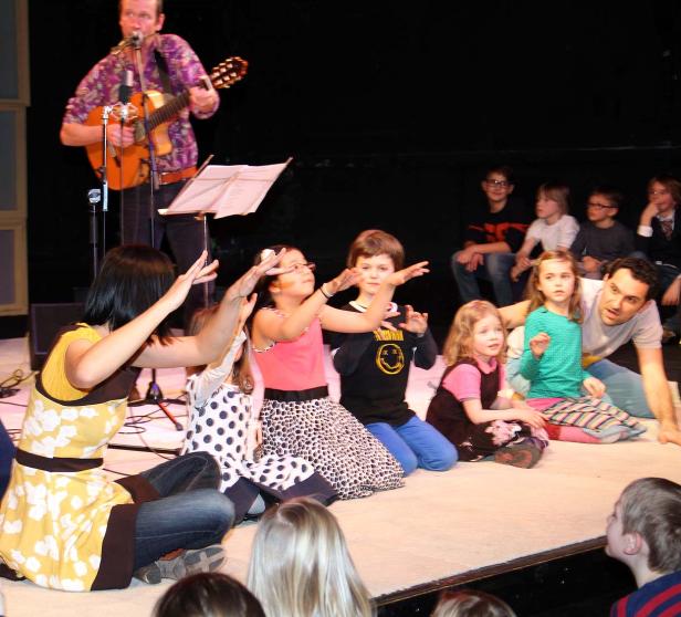 Ein Mann spielt Gitarre für eine Gruppe Kinder auf einer Bühne, die aufmerksam zusehen und gestikulieren.