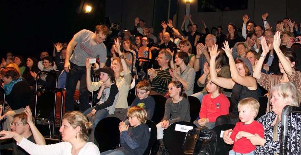 Ein Publikum sitzt in einem Theater und applaudiert oder winkt.