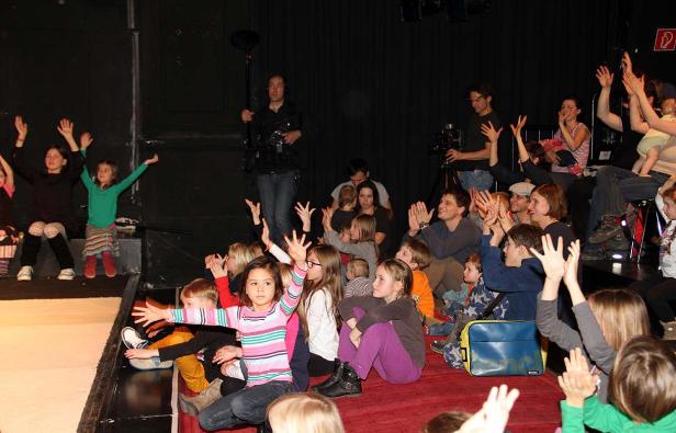 Eine Gruppe von Kindern und Erwachsenen sitzt auf einer Bühne und hebt die Hände.
