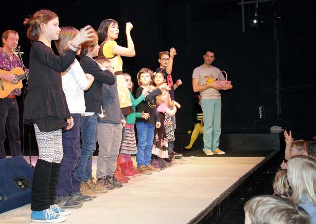 Eine Gruppe Kinder steht mit einer Frau und einem Mann auf einer Bühne.