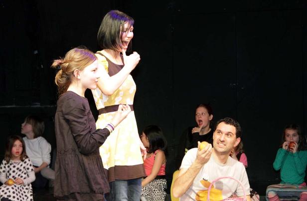 Eine Frau in einem gelben Kleid steht auf einer Bühne und gestikuliert, während andere Personen zusehen.