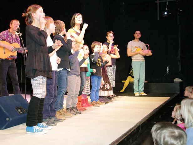 Eine Gruppe Kinder steht mit einem Mann mit Gitarre auf einer Bühne.
