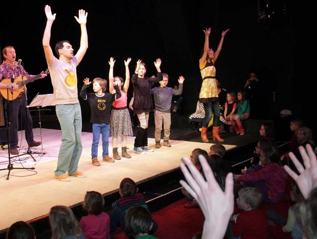 Eine Gruppe von Kindern und Erwachsenen steht auf einer Bühne und hebt die Arme.