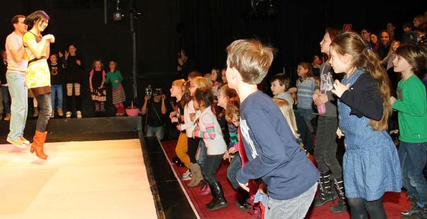 Eine Frau springt auf einer Bühne, während eine Gruppe Kinder zusieht.