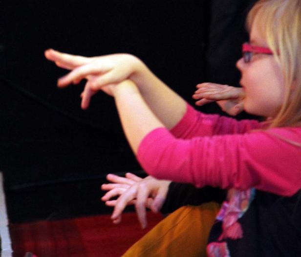 Ein Mädchen mit Brille macht Handbewegungen vor einem dunklen Hintergrund.