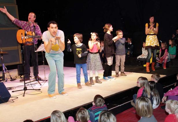 Eine Gruppe von Kindern steht mit zwei Erwachsenen auf einer Bühne vor einem Publikum.