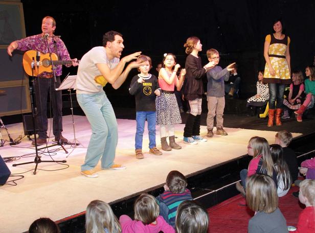 Ein Mann spielt Gitarre und ein anderer führt eine Gruppe Kinder auf einer Bühne an.