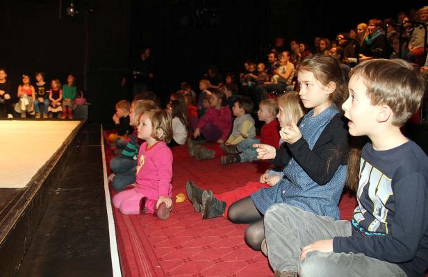 Eine Gruppe Kinder sitzt auf dem Boden und schaut aufmerksam auf eine Bühne.