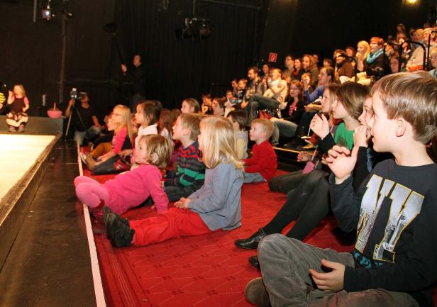 Kinder sitzen auf dem Boden und schauen aufmerksam auf eine Bühne.