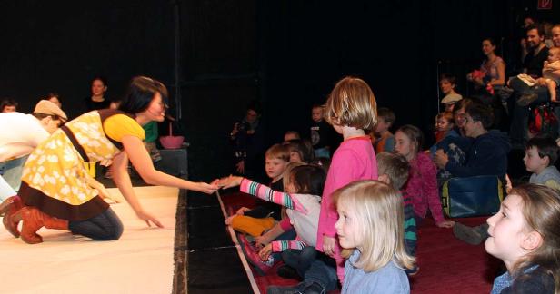 Eine Frau in einem gelben Kleid reicht einem Kind im Publikum die Hand.
