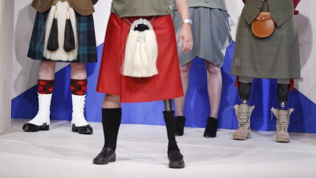 Mehrere Personen tragen Kilts und verschiedene Schuhe vor einer schottischen Flagge.