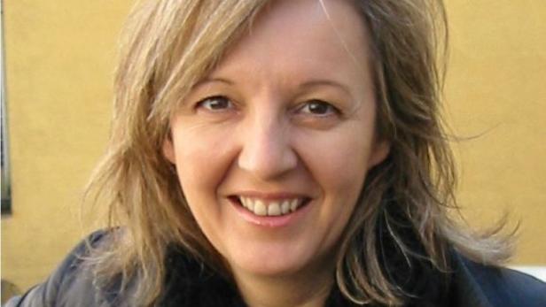 Eine lächelnde Frau mit blonden Haaren vor einem gelben Hintergrund.