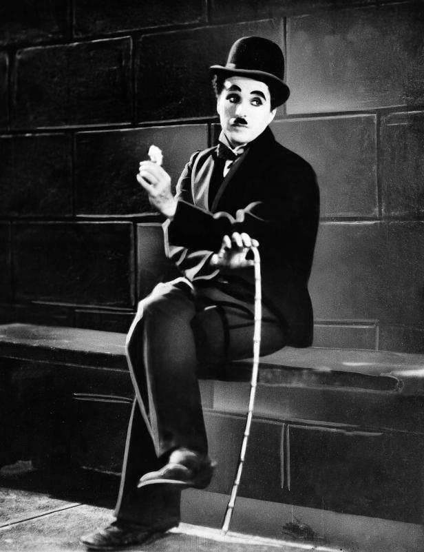 Charlie Chaplin sitzt mit Melone und Stock auf einer Bank.