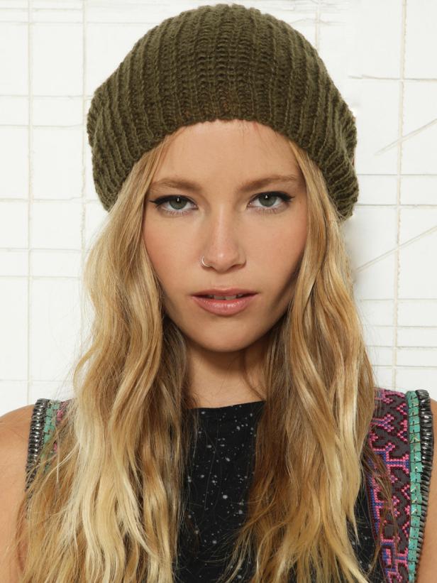 Eine Frau mit blonden, gewellten Haaren und einer olivgrünen Mütze posiert vor einer weißen Wand.