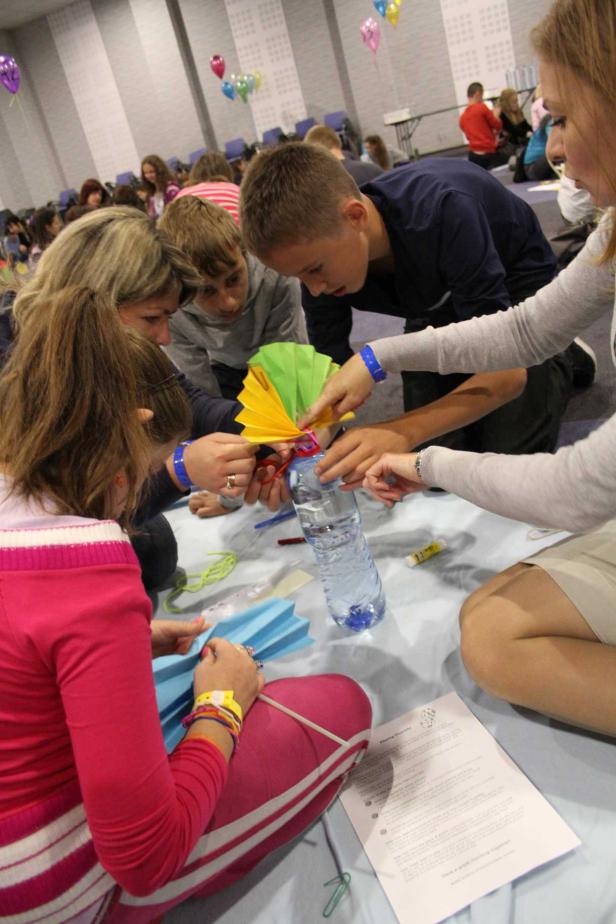 Eine Gruppe von Kindern und Erwachsenen bastelt gemeinsam an einem Tisch mit bunten Papierfächern und einer Wasserflasche.