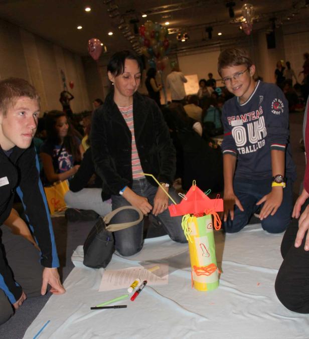 Eine Gruppe von Personen kniet um ein selbstgebasteltes Objekt aus Papier und Pappe.