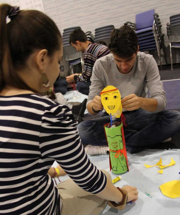 Drei Personen sitzen auf dem Boden und basteln an einer Superman-Figur aus Papier.