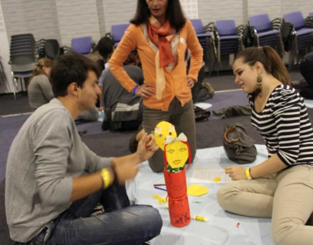 Eine Gruppe von Menschen sitzt auf dem Boden und bastelt an einer Puppe und Masken.