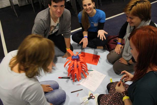 Eine Gruppe von Personen sitzt um ein Bastelprojekt mit einer Plastikflasche und rotem Papier.
