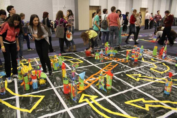 Eine Gruppe von Menschen betrachtet eine große Spielfläche mit bunten, selbstgemachten Figuren.