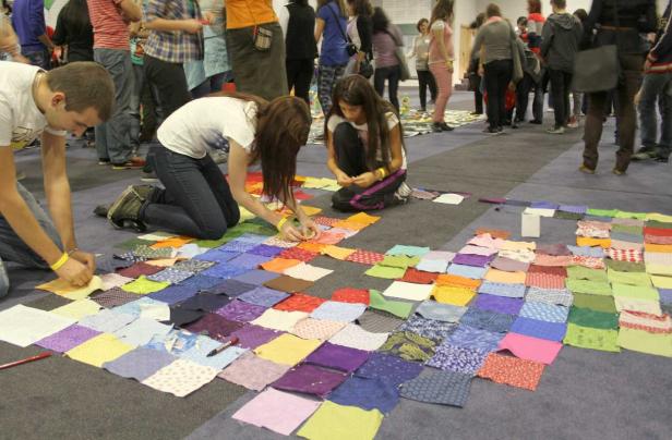Zwei junge Frauen ordnen bunte Stoffstücke auf dem Boden an, während andere Menschen im Hintergrund stehen.