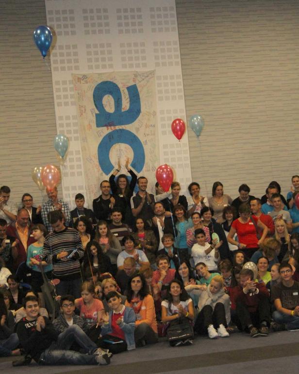 Eine große Gruppe von Menschen sitzt vor einem Banner und Luftballons.