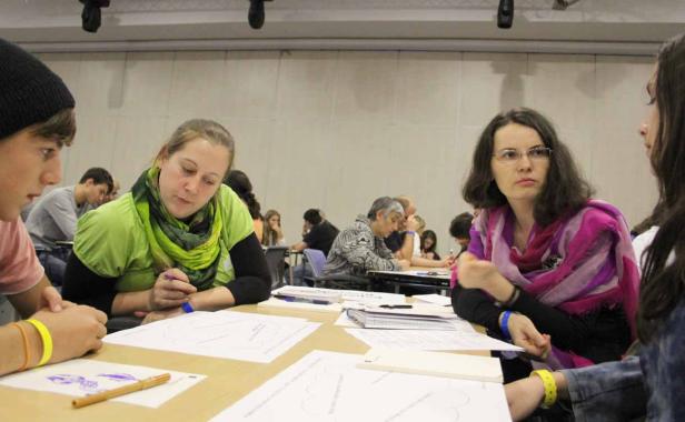 Mehrere Personen sitzen an Tischen und arbeiten an Aufgaben mit Stiften und Papier.