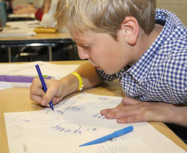 Ein Junge mit blonden Haaren zeichnet mit einem blauen Stift auf ein Blatt Papier.
