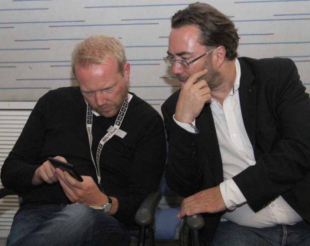 Zwei Männer sitzen nebeneinander; einer schaut auf ein Tablet, der andere denkt nach.