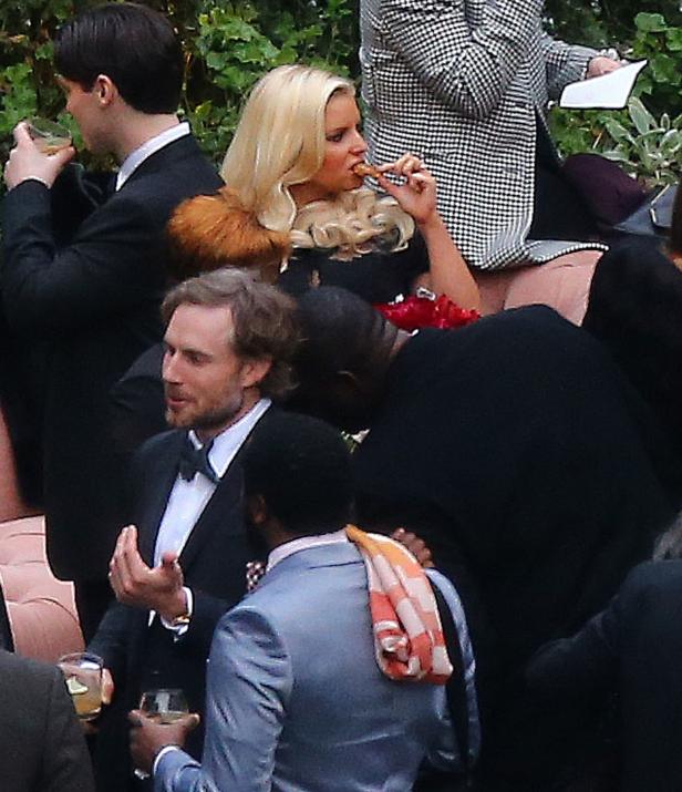 Eine blonde Frau isst inmitten einer Gruppe von Menschen bei einer Veranstaltung etwas.