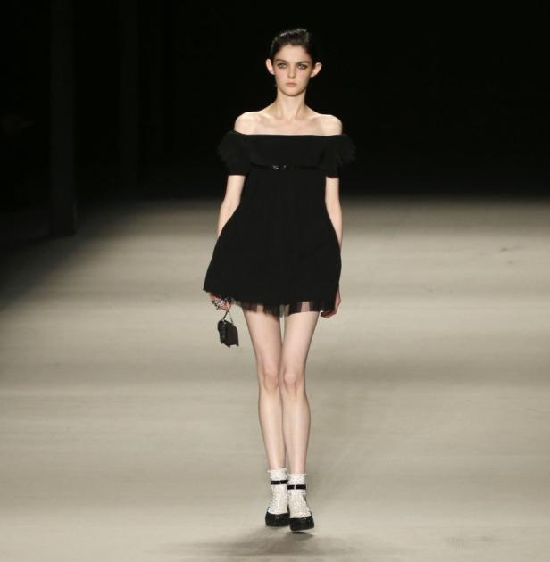 Ein Model präsentiert ein schwarzes, schulterfreies Kleid und auffällige Schuhe auf dem Laufsteg.