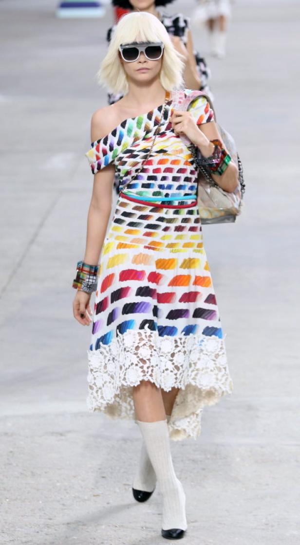 Eine Frau mit blondem Bob trägt ein farbenfrohes Kleid und eine Sonnenbrille auf dem Laufsteg.