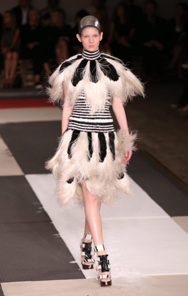 Ein Model mit silberner Kopfbedeckung präsentiert ein schwarz-weiß gestreiftes Kleid mit Federn auf dem Laufsteg.