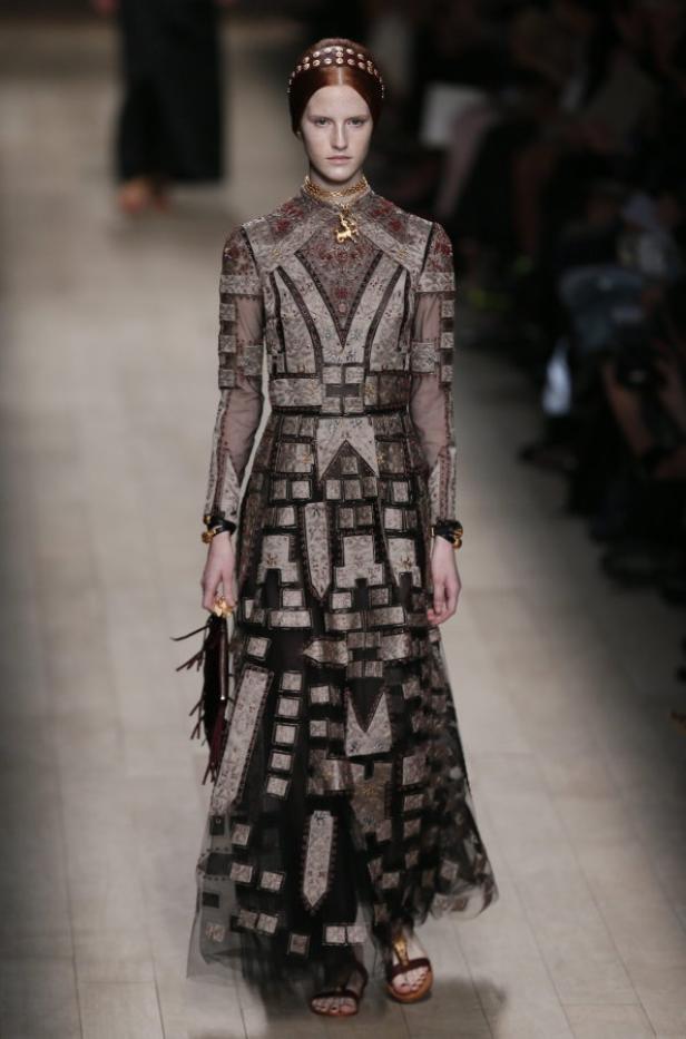 Ein Model präsentiert ein langes, gemustertes Kleid mit goldenem Schmuck auf einem Laufsteg.