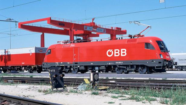 Eine rote ÖBB-Lokomotive zieht Güterwaggons unter einer Krananlage hindurch.